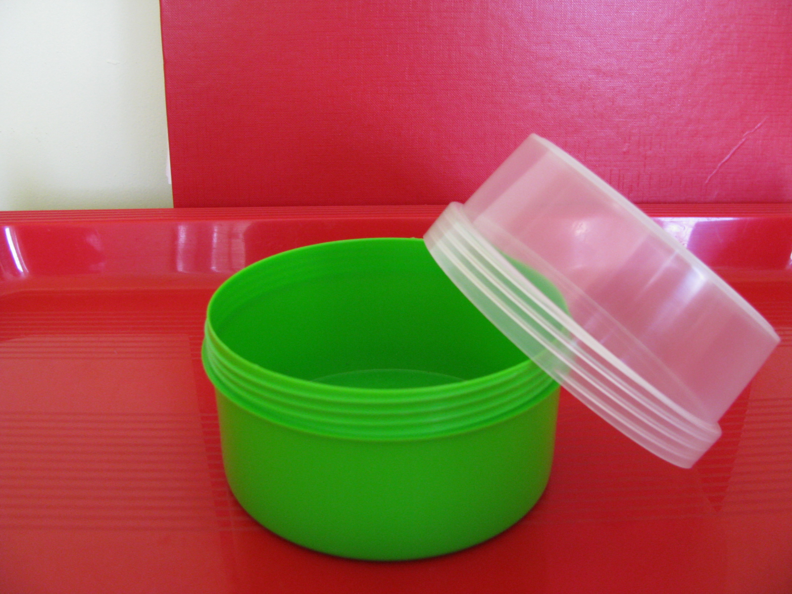 Otwarty, okrągły, zielony pojemnik z przezroczystą pokrywką na czerwonej tacy z czerwonym tłem.